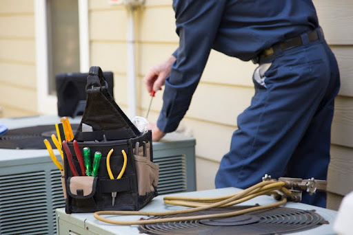 HVAC technician servicing an outdoor air conditioning unit next to a house. In the foreground is a black tool bag filled with pliers, screwdrivers, and other tools, resting on top of another HVAC unit with coiled hoses nearby.