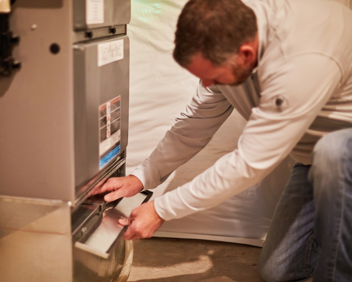 Allgood HVAC technician kneels beside a home furnace, sliding out the filter/access panel during installation.
