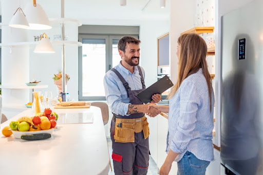 Smiling plumber wearing a tool belt and holding a clipboard shakes hands with a woman in a bright, modern kitchen. The scene suggests the completion of a successful home service appointment, with fresh fruit on the counter and natural light filling the space.